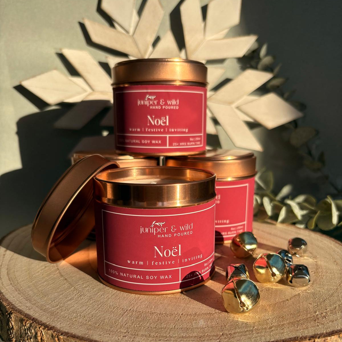 Three 'Noel' candles with red labels and bronze lids on a wooden surface with decorative elements.