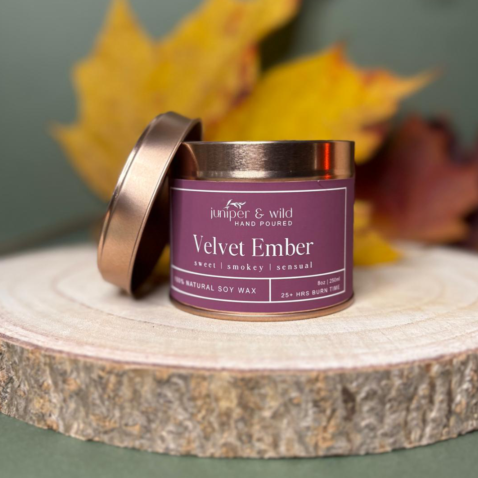 Candle tin labeled 'Velvet Ember' by Juniper & Wild on a wooden surface with autumn leaves in the background.