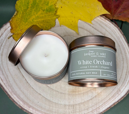 Two candles in metal tins with a 'White Orchard' label on a wooden surface with leaves.