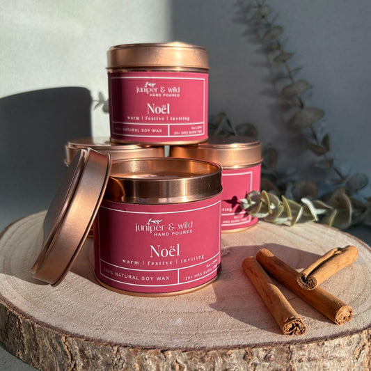 Three 'Noel' candles with copper lids on a wooden surface with cinnamon sticks.