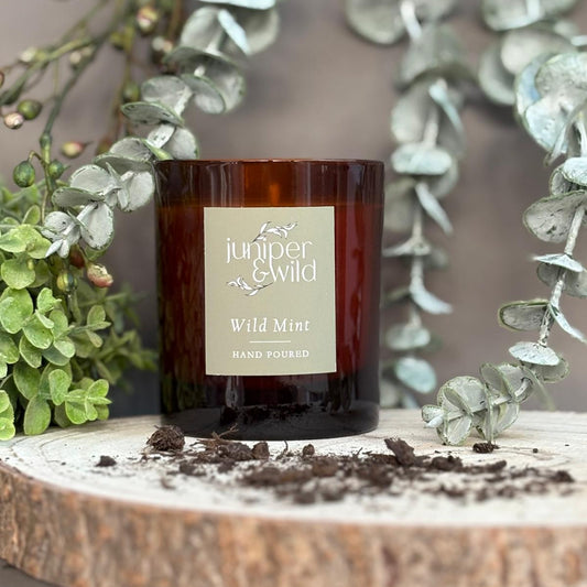 Candle labeled 'juniper & Wild' on a wooden surface with greenery in the background